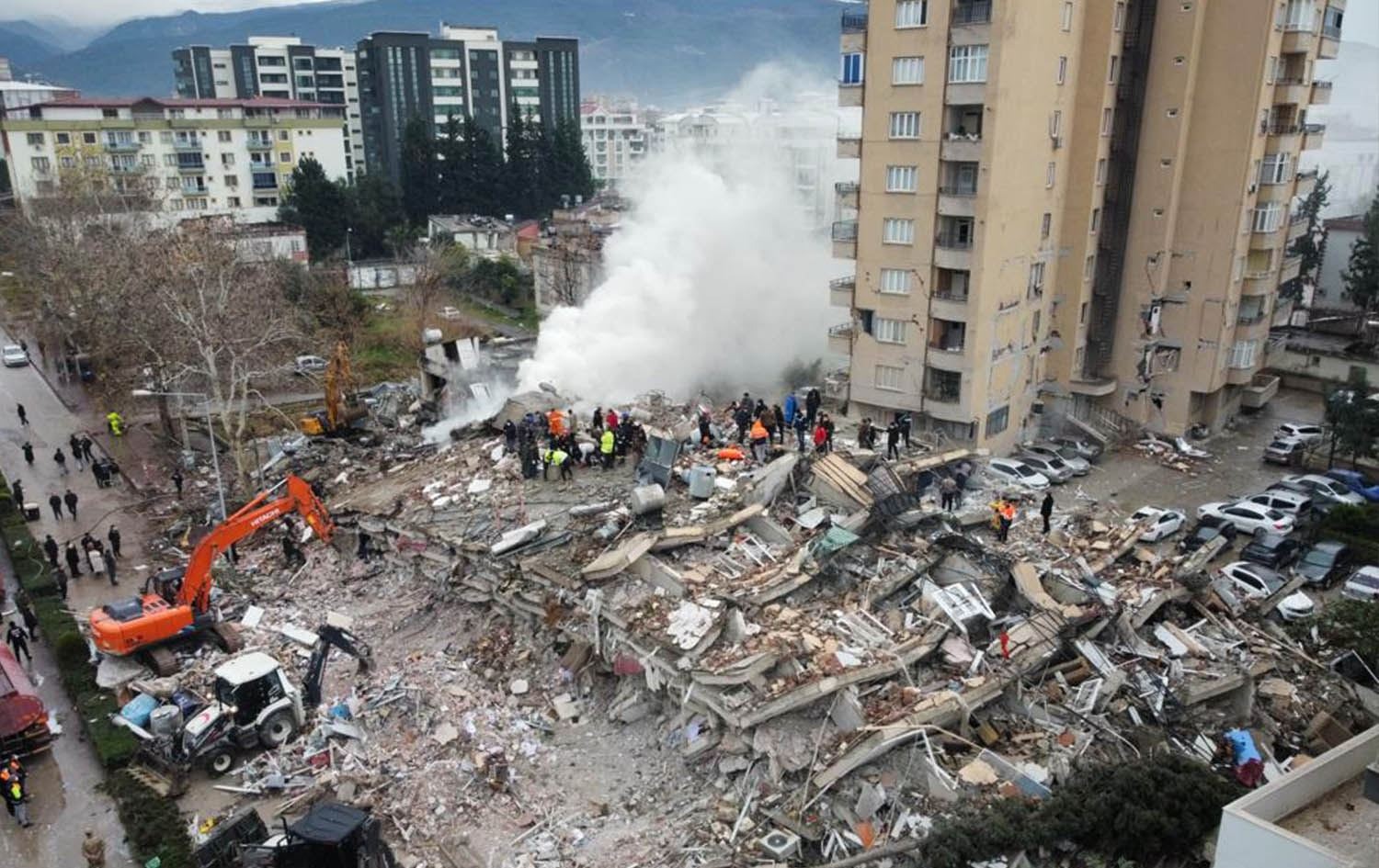 被災後の街の風景。手前の建物が倒壊しており、救助のゼッケンをつけた人が集まっている。左手前にショベルカー2台